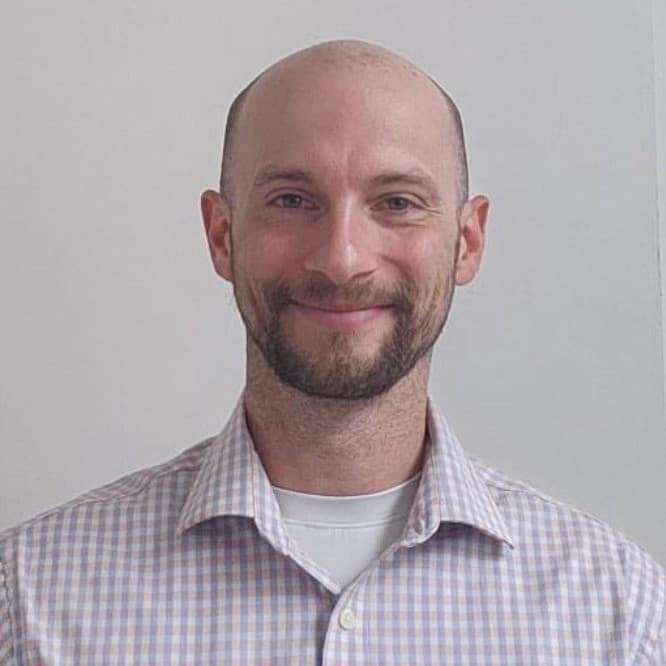 Smiling bald man with a beard and mustache wearing a light checkered button-down shirt.